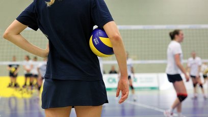 Eine Person im Vordergrund steht mit Rücken zur Kamera und hat einen Volleyball unter dem Arm. Im Hintergrund sieht man mehrere Personen in einem Hallen-Volleyballfeld spielen.