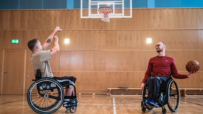 2 Personen im Rollstuhl spielen gemeinsam Basketball in einer Turnhalle